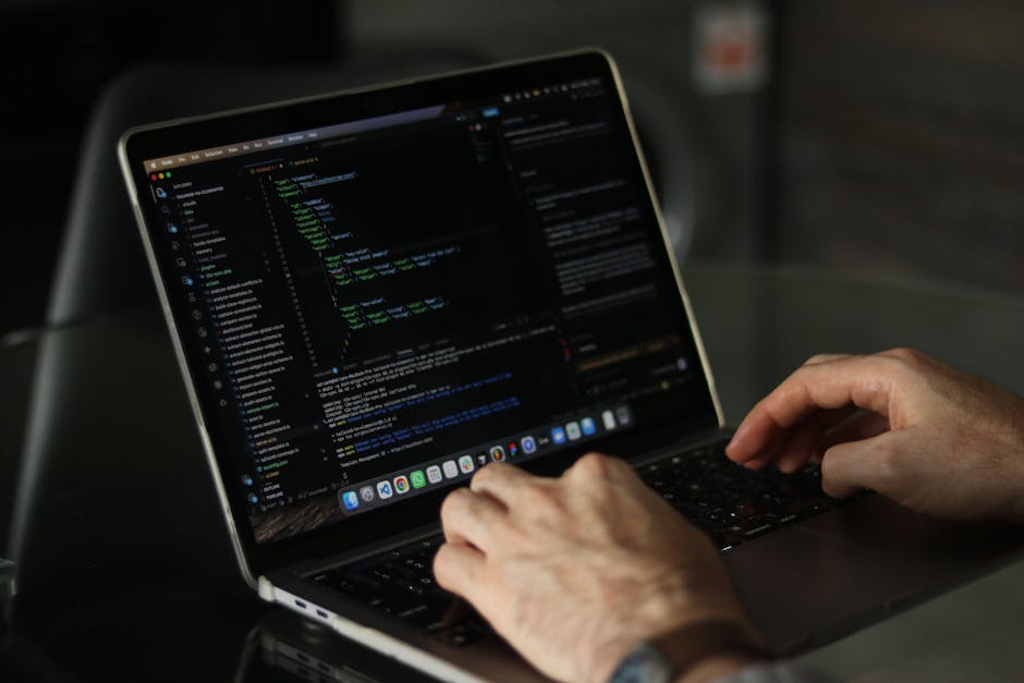 A person working on a laptop with a dark screen displaying lines of code in a text editor, sitting at a desk with a reflective black surface. The individual's hands are visible, with fingers poised ov
